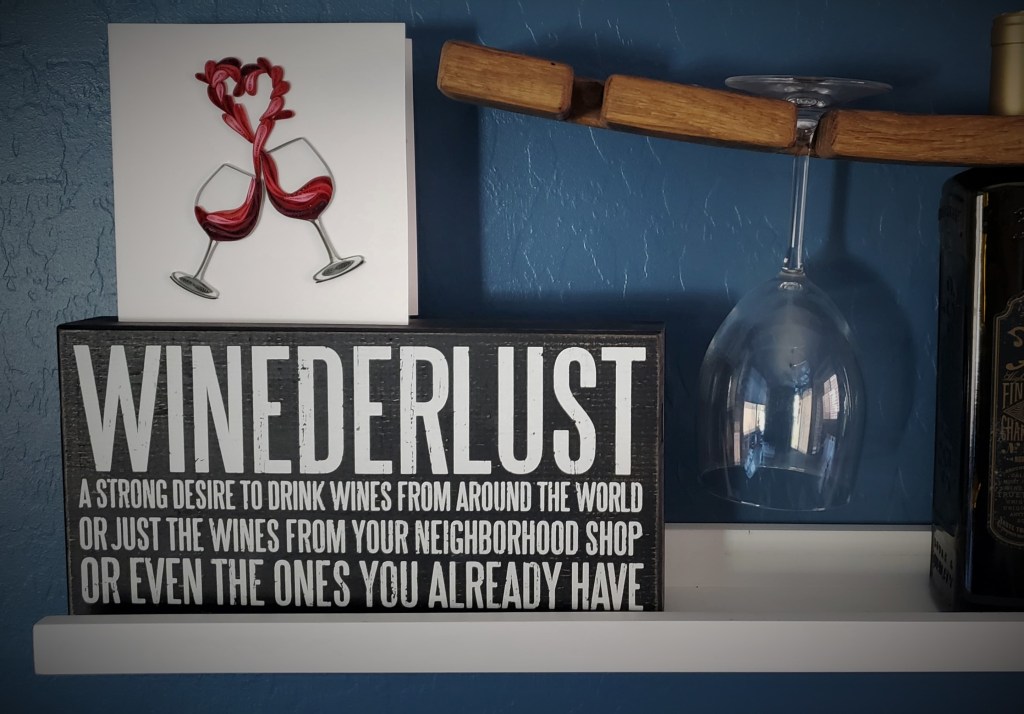 Sign on a wall: "Winederlust - a strong desire to drink wines form around the world or just the wines from your neighborhood shop or even the ones you already have." Photo also shows a decorative card with wine glasses and wine shaped in the shape of a heart.