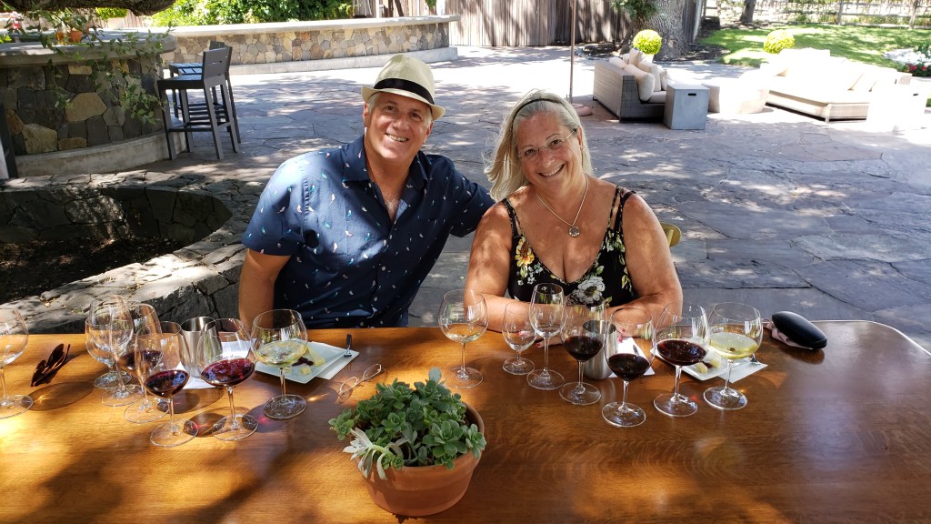 couple smiling sitting at a table set for a wine tasting under a shade tree
